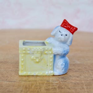 May include: A small, vintage ceramic planter featuring a light blue dog wearing a red bow, leaning against a yellow chest. The planter has a rectangular opening for plants. The dog is detailed with black eyes and a small nose. The chest has a decorative clasp.