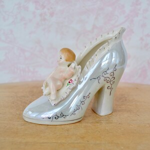 May include: A pearlescent ceramic shoe-shaped figurine with a high heel, featuring a small cherub figure resting on a leaf-shaped base. The shoe has gold trim and decorative swirls.
