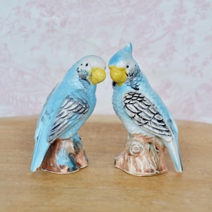 May include: Two ceramic parakeet figurines. The birds are light blue with white heads and yellow beaks. They are perched on brown tree trunk bases. The wings have gray and white detailing. The background is a light pink floral pattern.