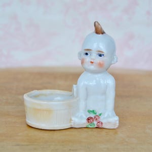 May include: A white porcelain figurine of a baby with a small tub. The baby has blue eyes, rosy cheeks, and a brown topknot. The tub is light beige. A cluster of red and green flowers is at the baby's feet.