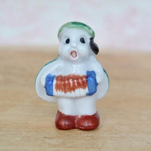 May include: A small porcelain figurine of a person wearing a green hat and holding a red and blue accordion. The figurine is painted with white, red, blue, and green.