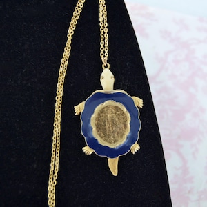 May include: A gold-tone necklace featuring a turtle-shaped pendant. The pendant has a cream-colored head and limbs, with a dark blue enamel border surrounding a textured, gold-colored center shell. The chain is a thin, matching gold-tone metal link.
