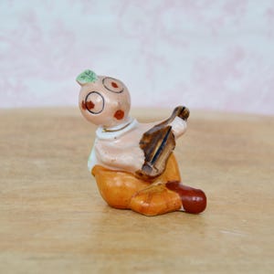 May include: A small, vintage ceramic figurine of a seated figure playing a stringed instrument. The figurine has a peach-colored face with large eyes and rosy cheeks, wearing orange pants and a brown instrument.