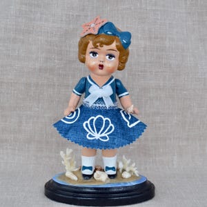 May include: A vintage-style figurine of a girl in a blue sailor dress with a white bow and seashell design. The doll has a bow and starfish in her hair and stands on a base with seashells and coral.