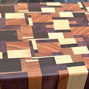 Puede incluir: Una tabla de cortar rectangular de madera con un patrón de mosaico de varios tonos de madera, incluyendo beige claro, marrón medio, marrón oscuro y morado. La tabla mide aproximadamente 46 cm de largo y 30 cm de ancho.