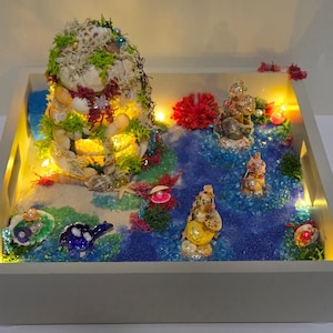 Könnte beinhalten: Dekorative Tablett mit einer Miniatur-Ozeanszene, einschließlich eines Muschelturms, Meerjungfrau-Figuren und einem blau-weißen Wassereffekt. Das Tablett wird mit kleinen Lichtern beleuchtet, wodurch eine skurrile Darstellung entsteht.