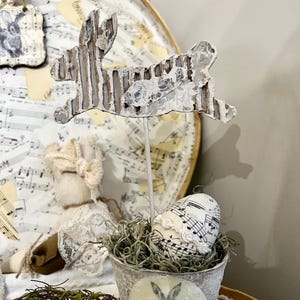 May include: A white ceramic bunny cutout with a distressed finish, perched on a stick, is displayed in a small metal bucket filled with green moss. A white egg with a musical note design is nestled in the moss. The bucket has a vintage bunny design on the side.