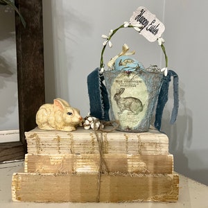 May include: A white ceramic bunny figurine sits on a stack of vintage books. A blue and white fabric basket with a floral design and the text "Belle Feronniere" is filled with faux flowers and eggs. A white tag with the text "Happy Easter" is attached to the basket.