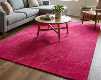 May include: A large, rectangular, solid magenta area rug with a short, dense pile texture. The rug is positioned on a wooden floor in a living room setting, extending underneath a round wooden coffee table, a gray sectional sofa, and a chair.