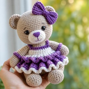 May include: A beige crocheted teddy bear featuring a purple bow on its head and a purple nose. The bear wears a white and purple crocheted dress with a ruffled skirt and white trim. The toy has black safety eyes and a stitched mouth.