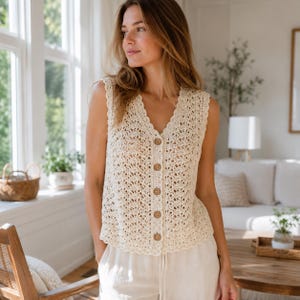 May include: A sleeveless cream-colored crochet vest featuring a V-neckline and a front button-down closure with five round wooden buttons. The garment is crafted with an intricate openwork lace pattern and finished with a scalloped edge trim.