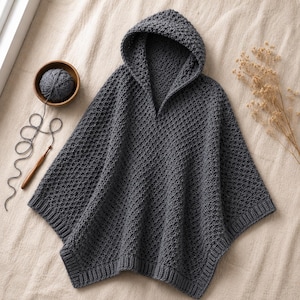 May include: A dark gray hooded poncho featuring a textured, repeating stitch pattern throughout the body. The garment includes a ribbed hem along the bottom edge and a hood. A ball of matching gray yarn and a wooden crochet hook are visible nearby.