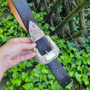 Może przedstawiać: Czarny pasek z teksturowanym wzorem i ozdobną srebrną klamrą. Klamra ma bogaty wzór z kwiatowymi i geometrycznymi detalami. Wnętrze paska jest w kolorze jasnobrązowym.