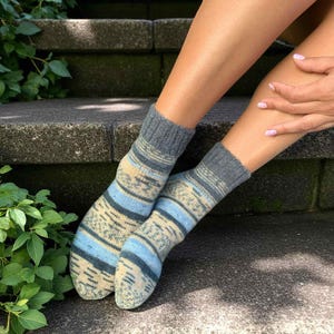 Könnte beinhalten: Ein Paar gemusterte Socken in Blau-, Beige- und Grautönen. Die Socken haben Streifen und geometrische Muster. Die gerippten Bündchen sind dunkelgrau. Die Socken werden an den Füßen einer Person getragen.