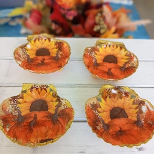 May include: Four decorative seashell-shaped dishes. Each dish features a vibrant design of a sunflower and pumpkins in shades of orange, yellow, and brown. The edges of the dishes have a gold trim, and the background is white.
