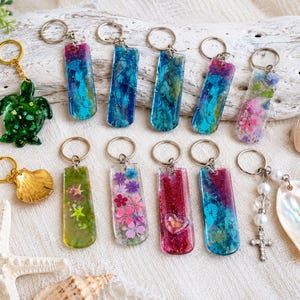May include: A collection of resin keychains featuring various designs including a green sea turtle, rectangular shapes with blue, pink, and glittery swirls, pressed flowers, a heart accent, a gold seashell, and a cross charm with white beads.