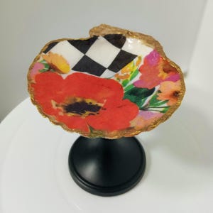 May include: A decorative seashell-shaped trinket dish with a black base. The dish features a vibrant floral and checkered pattern with a large red poppy. The edges are trimmed with gold.