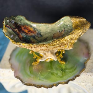 May include: A decorative oyster shell bowl with gold-leafed edges, featuring an interior painting of a fox. The shell rests on a gold-toned metal stand, which is positioned atop a circular, translucent green and white resin coaster with gold trim.