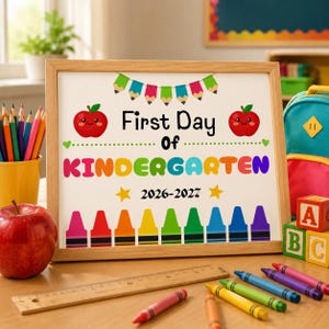 Puede incluir: Un letrero con marco de madera con el texto First Day Of KINDERGARTEN 2026-2027. El diseño presenta banderines de lápices coloridos, dos manzanas rojas sonrientes y una fila de crayones multicolores en la parte inferior sobre fondo blanco.