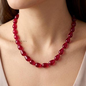 Könnte beinhalten: Eine Halskette aus tropfenförmigen, tiefroten Perlen. Die Perlen sind zu einer Halskette aufgereiht. Der Text "TES GEMS" ist auf der linken Seite des Bildes zu sehen. Die Halskette wird um den Hals getragen.