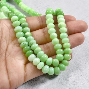 May include: A close-up of strands of light green gemstone beads. The beads are oval-shaped and have a smooth, polished surface. The beads are held in a hand. The text "TESLAGEMS" is visible in the image.