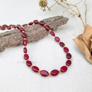 May include: A necklace featuring oval-shaped, deep red gemstones strung together with small gold-colored beads. The necklace is displayed on a light-colored surface with a piece of wood and dried foliage in the background. The text "TESLA GEMS" is visible.