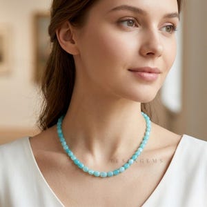 May include: A light blue beaded necklace with rounded beads. The necklace is worn around the neck. The text "TESLA GEMS" is visible on the right side of the necklace. The person is wearing a white top.