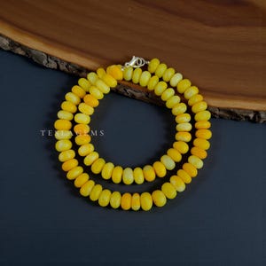 Puede incluir: Un collar hecho de cuentas de piedras preciosas amarillas. Las cuentas son de forma ovalada y están ensartadas, formando una doble hebra. El collar tiene un cierre plateado. La marca "TESLAGEMS" es visible.