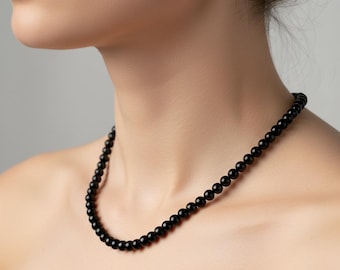 Puede incluir: Un collar de cuentas negras con pequeñas cuentas redondas. El collar se lleva alrededor del cuello. La imagen está sobre un fondo neutro. La palabra "TESLAGEMS" es visible en la esquina inferior izquierda.