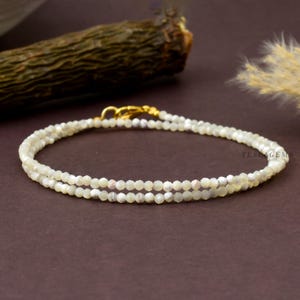 Könnte beinhalten: Ein zartes, zweireihiges Armband aus kleinen, facettierten, cremeweißen Perlen mit einem dezenten Perlmuttschimmer. Das Armband verfügt über einen goldfarbenen Karabinerverschluss und enthält den Text TESLAGEMS in der Nähe des Verschlusses.