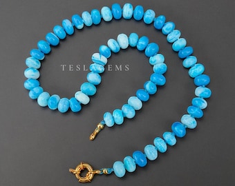 Op de afbeelding: Een ketting bestaande uit lichtblauwe en witte ovaalvormige kralen. De kralen zijn aan elkaar geregen en vormen een gebogen vorm. De ketting heeft een goudkleurige sluiting en een klein goudkleurig accent in het midden. De tekst "TESLA GEMS" is zichtbaar.