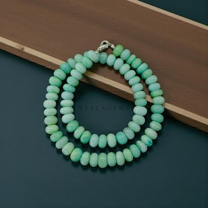 May include: A double-strand necklace featuring light green, oval-shaped beads. The beads are strung together with a silver clasp. The necklace is displayed on a dark surface with a wooden accent. The text "TESLAGEMS" is visible.