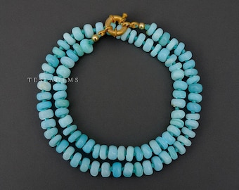 Op de afbeelding: Een ketting met drie strengen lichtblauwe, ronde kralen. De sluiting is goudkleurig. De tekst "TESLA GEMS" is links in de afbeelding zichtbaar. De ketting is afgebeeld tegen een donkergrijze achtergrond. De kralen zijn ongeveer 0,5 cm in diameter.