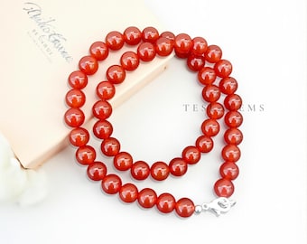 May include: A necklace composed of polished, round, red-orange beads. The beads are strung together, forming a double strand that fastens with a silver clasp. The necklace is displayed on a white surface with a light-colored box in the background.