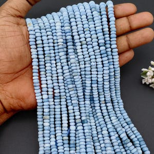 May include: Strands of light blue gemstone beads. The beads are small, cylindrical, and have a slightly irregular shape. The color is a soft, muted blue, with variations in tone. The beads are strung together, creating a flowing, textured appearance. The beads are held in a hand.