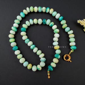May include: A beaded necklace featuring oval-shaped gemstones in shades of white, green, and yellow. The necklace has a gold-colored clasp and the text "TES GEMS" is visible. The necklace is displayed on a dark background.
