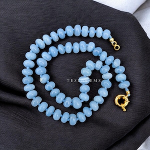 May include: A light blue beaded necklace with a gold clasp. The necklace is composed of small, rounded beads strung together on a light blue cord. The text "TEST GEMS" is visible on the background.