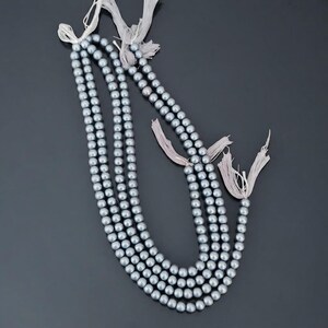 May include: A long strand of round, metallic grey beads with a smooth, reflective finish. The strand is looped and features light-coloured, frayed fabric tassels at the ends, creating a continuous, decorative chain of uniform, spherical components.