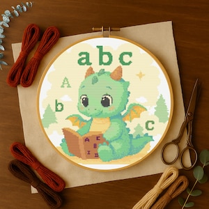 Pode incluir: Bastidor de bordado em ponto cruz com um dragão verde lendo um livro com as letras A, B e C. O dragão tem chifres e asas laranja. O fundo é amarelo com árvores e nuvens. Fios de bordar e tesouras também são visíveis.