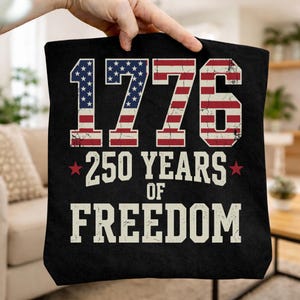 May include: Black tote bag featuring the text "1776 250 YEARS OF FREEDOM" in white and red, white, and blue. The numbers in "1776" are designed to look like the American flag. The bag is held up by a hand.