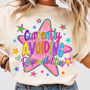 May include: A cream-colored t-shirt featuring a large pink star graphic with a glittery texture. The text "Currently AVOIDING Responsibilities" is printed in colorful, bold letters over the star, surrounded by smaller multi-colored stars.
