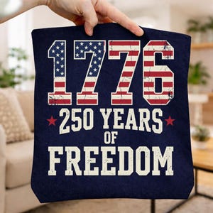 May include: A navy blue tote bag featuring the text "1776 250 YEARS OF FREEDOM" in white. The numbers and letters are designed with the American flag's stars and stripes. Red stars flank "250 YEARS".