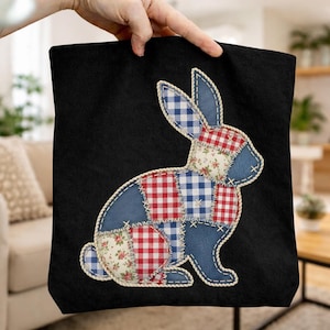 Può includere: Borsa tote nera con un design a coniglio patchwork. Il coniglio è realizzato in tessuto blu, a quadri rossi e con motivi floreali. Il coniglio è delineato con un punto bianco. La borsa tote è tenuta da una mano.