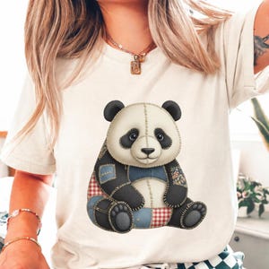 May include: Cream-colored t-shirt featuring a seated panda bear design. The panda has a patchwork appearance with black, white, red, and blue fabric sections. The panda's eyes are black, and it has black ears and feet.