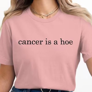 May include: A light pink t-shirt with the phrase "cancer is a hoe" printed in black, centered on the front. The shirt has a crew neck and short sleeves. A gold necklace is visible at the neckline.