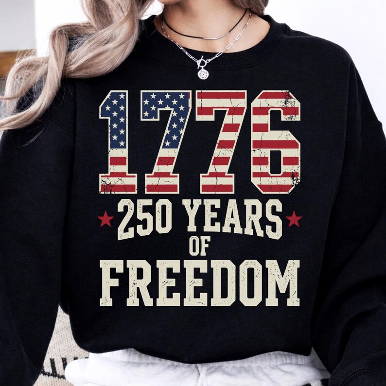 May include: Black sweatshirt featuring the text "1776 250 YEARS OF FREEDOM". The numbers and letters are white, with the numbers 1776 designed to resemble the American flag. Two red stars flank the words "250 YEARS".