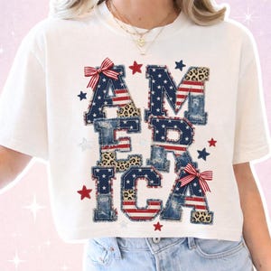 May include: White crop top featuring the word "AMERICA" in a patchwork design. The letters incorporate denim, leopard print, and the American flag. Red and blue stars and bows add to the patriotic theme.