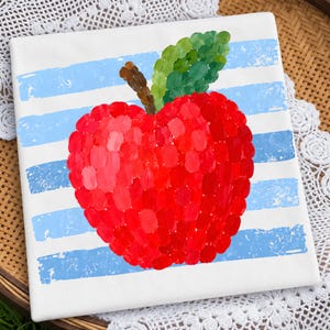 Peut inclure: Un carreau carré blanc orné d'une pomme rouge peinte avec une feuille verte et une tige brune. La pomme est composée de coups de pinceau visibles et centrée sur un fond de rayures horizontales bleu clair à la finition texturée et usée.