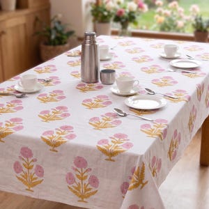 May include: A white tablecloth featuring a repeating pattern of pink flowers with yellow stems and leaves. The table is set with white ceramic cups, saucers, and plates, along with silver spoons and a tall stainless steel vacuum insulated thermos.