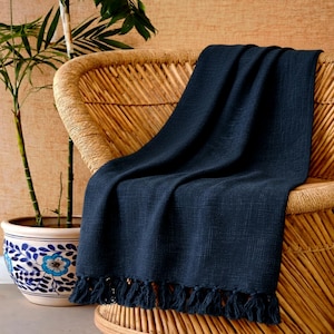Puede incluir: Una manta tejida azul marino con flecos, drapeada sobre una silla de mimbre. La manta tiene una apariencia texturizada y se presenta en un entorno de decoración del hogar. Una planta en maceta es visible en el fondo.